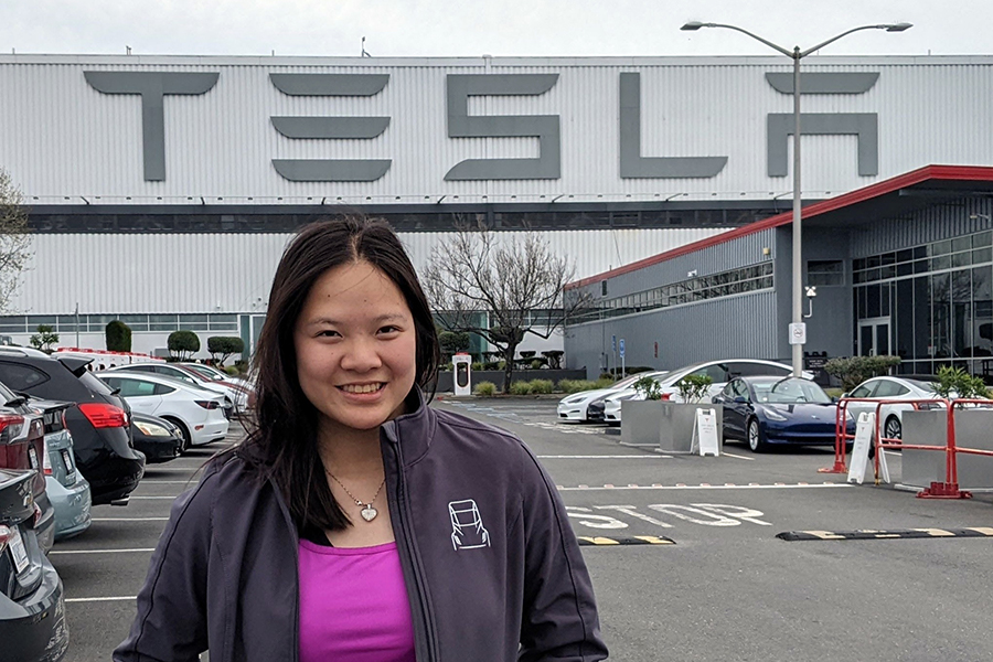 Hana is wearing a pink shirt and a gray jacket. She is standing in front of a Tesla facility.