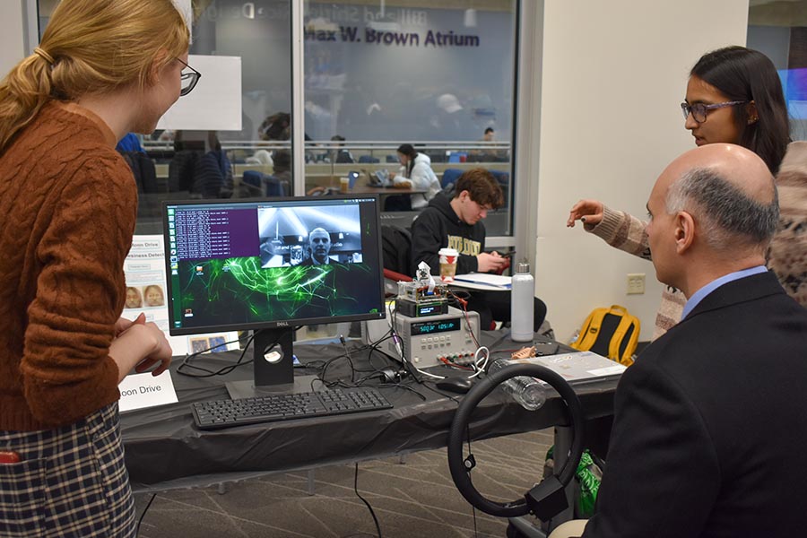 Two female students, a team from the Fall 2022 Spark Challenge, explains their project to Dimitrios Peroulis, Michael and Katherine Birck Head and Reilly Professor of ECE, who is seated at computer that also has an attached apparatus