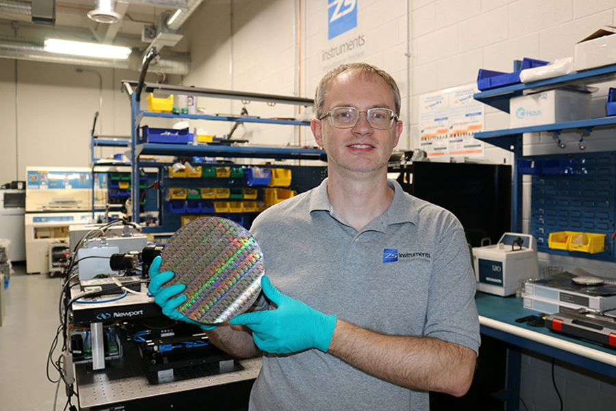 Sergey Zakharov is holding a wafer in a laboratory setting. He is wearing a gray polo shirt with safety glasses and gloves. Behind him are various pieces of lab equipment.