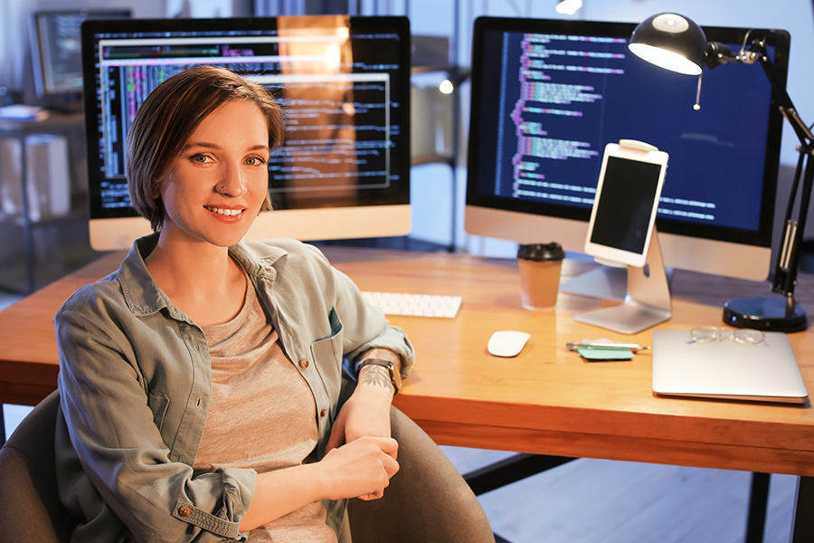 A young woman sits in front of a two computer screens with lots of code on them.