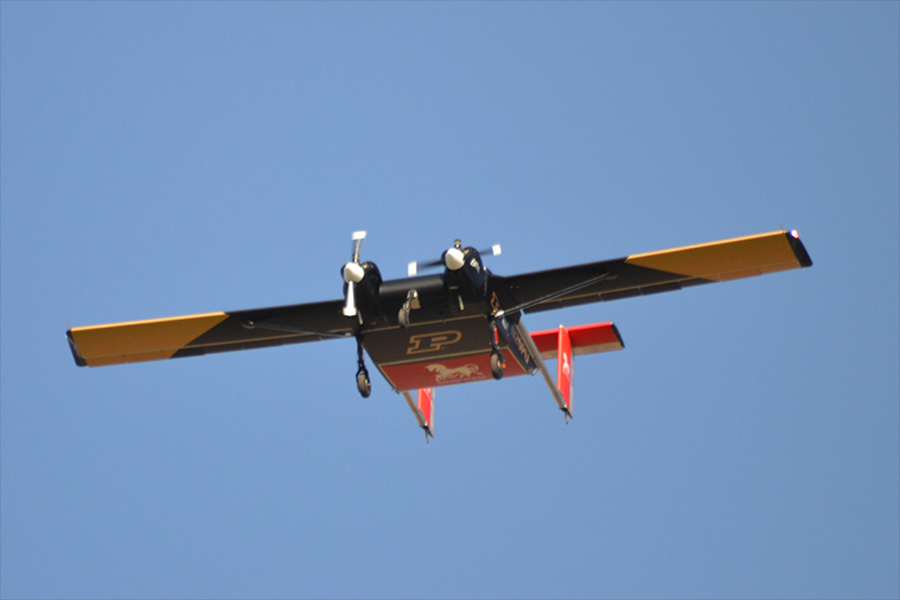 The Windracers ULTRA is flying in a clear blue sky. The craft is painted black and gold and has the Purdue P painted on its underside.