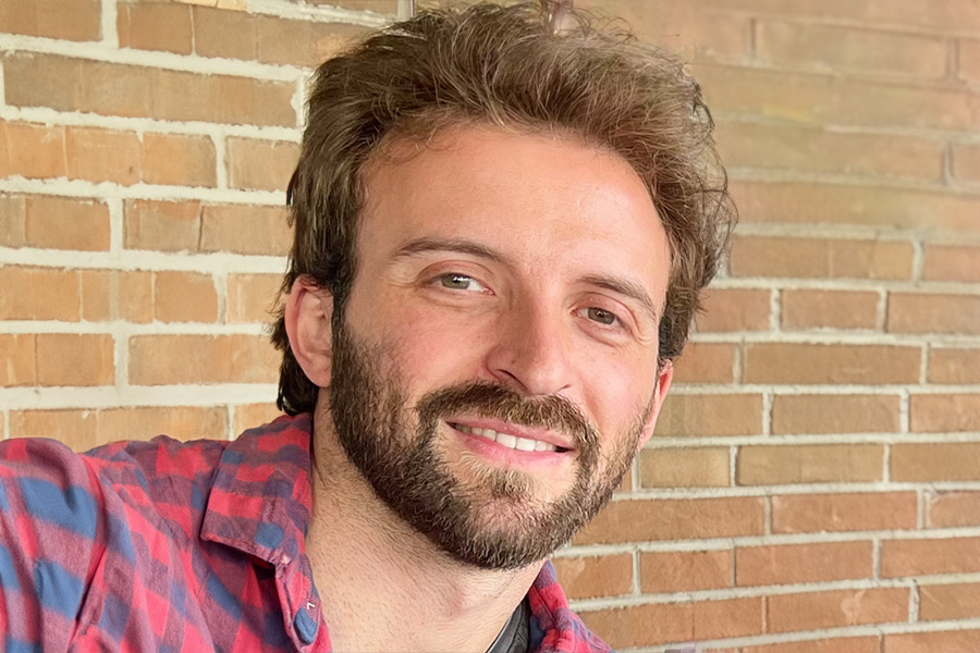 A bearded man wearing a red and blue shirt, standing infront of a brick wall, smiling and looking directly at the camera.