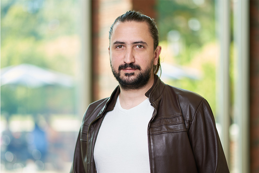 A bearded man wearing a leather jacket poses in front of a large window