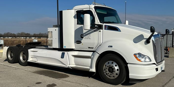 The semi truck equipped with the Cummins technology is parked on the side of a street with a fence. The truck is painted white.