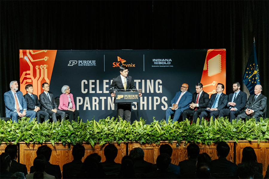 Indiana state leaders, Purdue leaders, and SK hynix leaders sit on a stage. Behind them is a banner that reads 'celebrating partnerships'. Purdue President Mung Chiang is speaking at a podium.