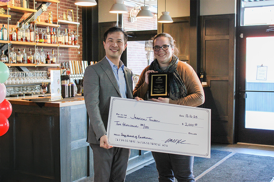 A smiling man in a suit and a woman holding a plaque pose with a large check. They stand in a warmly lit bar with shelves full of bottles.