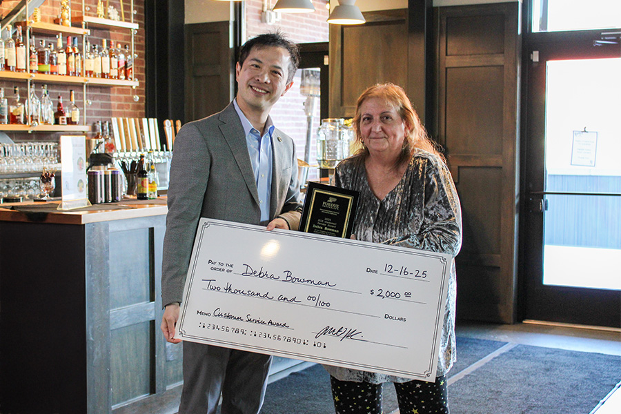 Man in a suit and woman holding an award plaque smile while posing with a large mock check for $2,000. They are standing in a dimly lit bar.