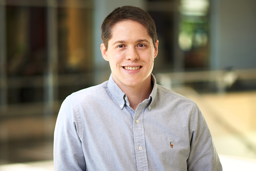 Smiling person in a blue button-up shirt stands in a softly lit indoor setting, conveying a warm and friendly tone. Blurred background.