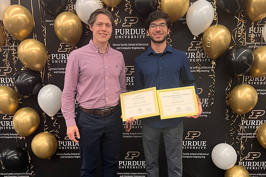 A Purdue ECE professor and a Purdue ECE student pose at an awards event in front of a black Purdue University backdrop with gold, black, and white balloons; one student holds framed certificates for Austin Gayte and Arjun Gupte.