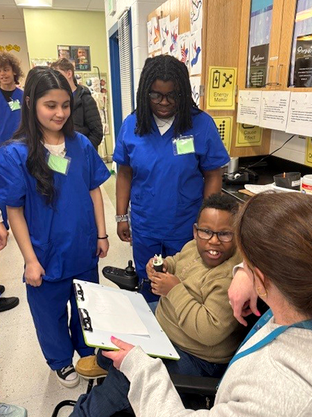 two students in blue scrubs with boy in wheelchair and woman
