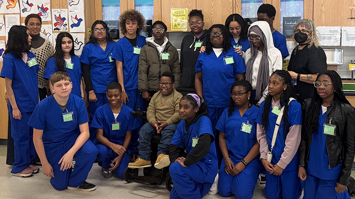 group of seventh grade EPICS students in blue scrubs with teachers and boy in wheelchair