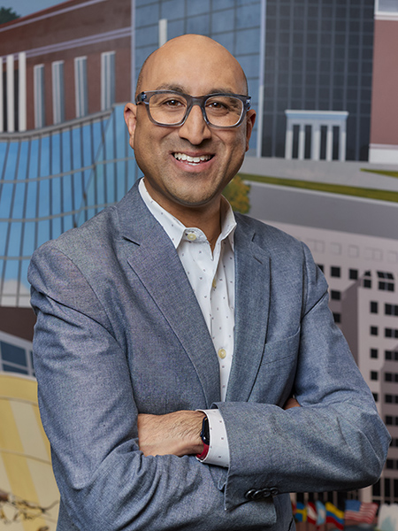 Man with blue-rimmed glasses, bald, smiling wearing gray suit coat and patterned button-up shirt