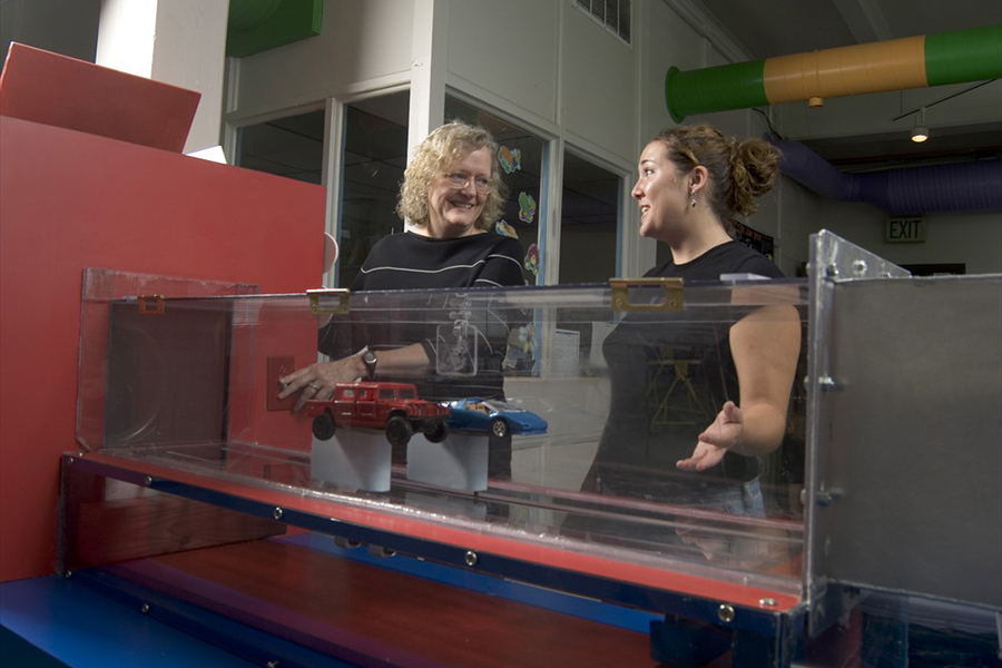 Two women standing behind an EPICS project, tunnel with cars in tube