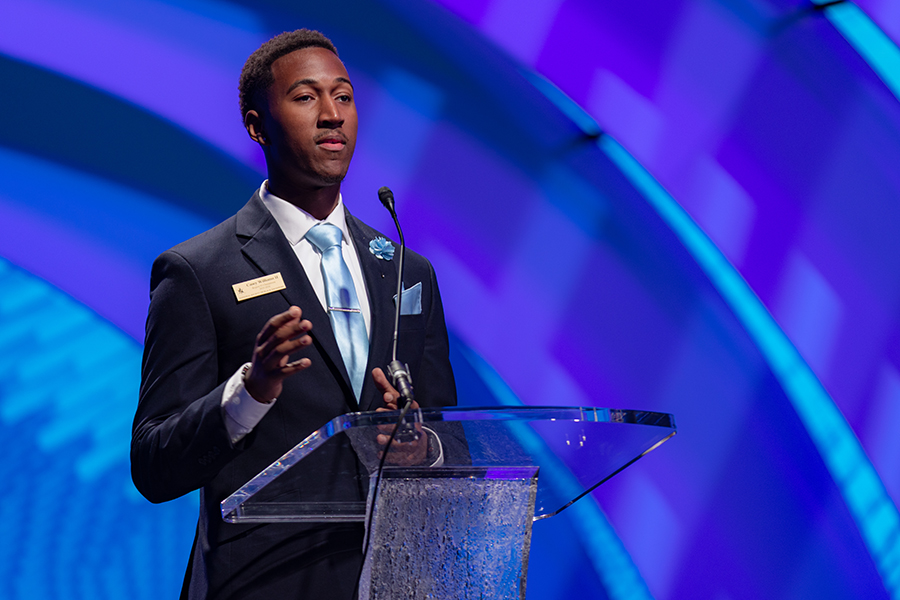 Student wearing a dark suit with a light blue tie, pocket square, standing on a stage and giving a speech