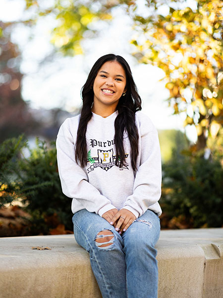 Student with long dark hair, smiling, wearing a Purdue sweatshirt and ripped jeans
